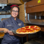 Kelly Potter holding a freshly baked pepperoni pizza in front of a commercial oven at Prohibition Pizza